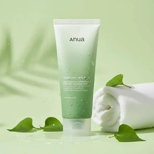 Un tube de mousse nettoyante pour le visage Anua Heartleaf + BHA est posé debout sur une surface verte, accompagné de feuilles de cœur fraîches et d’une serviette blanche roulée, le tout sur fond vert clair avec des gouttelettes d’eau.