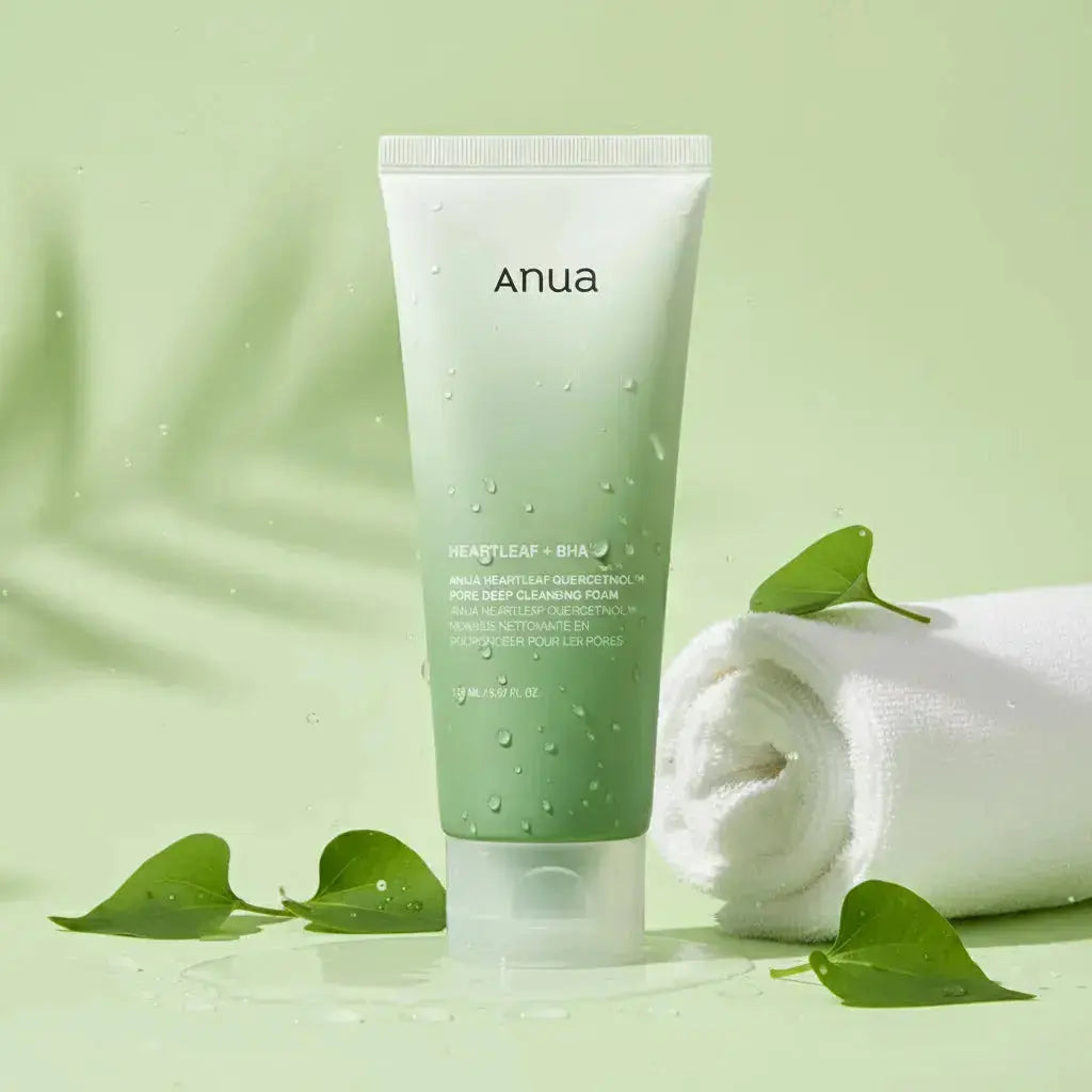 Un tube de mousse nettoyante pour le visage Anua Heartleaf + BHA est posé debout sur une surface verte, accompagné de feuilles de cœur fraîches et d’une serviette blanche roulée, le tout sur fond vert clair avec des gouttelettes d’eau.